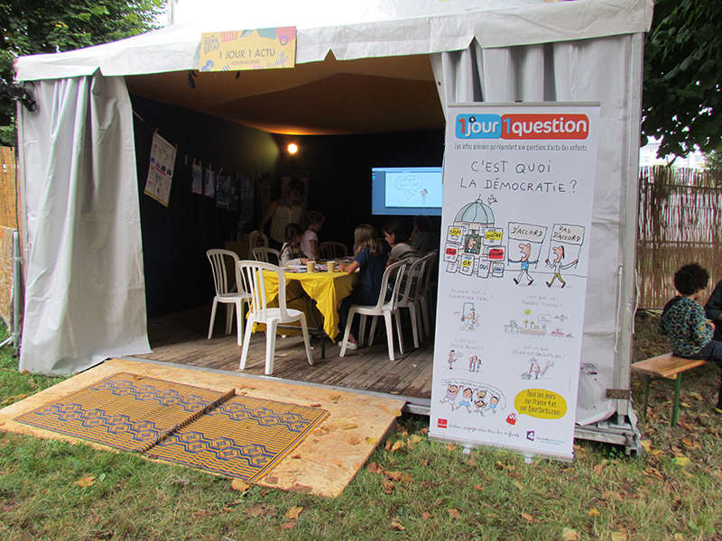 Une tente à l'intérieur de laquelle des enfants sont assis autour d'une table pour écrire et dessiner la prochaine vidéo d'1jour1question