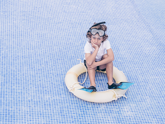 Un enfant avec masque, tuba, palmes et bouée, l'air désolé de se retrouver dans une piscine vide. 