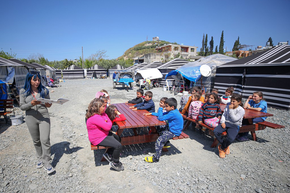 Une photo d'une femme proposant des activités en extérieur à des enfants de tous âges. Les enfants sont assis à des tables de pique-nique. On voit des tentes d'habitation autour.