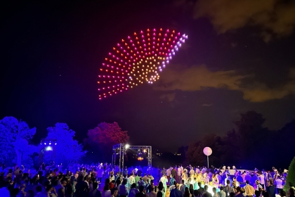 Image extraite d'un spectacle de drones qui forment comme un éventail rouge, rose et jaune dans le ciel noir.