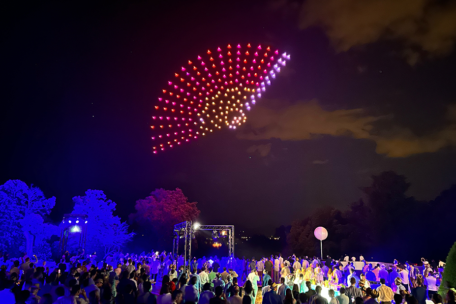 Image extraite d'un spectacle de drones qui forment comme un éventail rouge, rose et jaune dans le ciel noir.