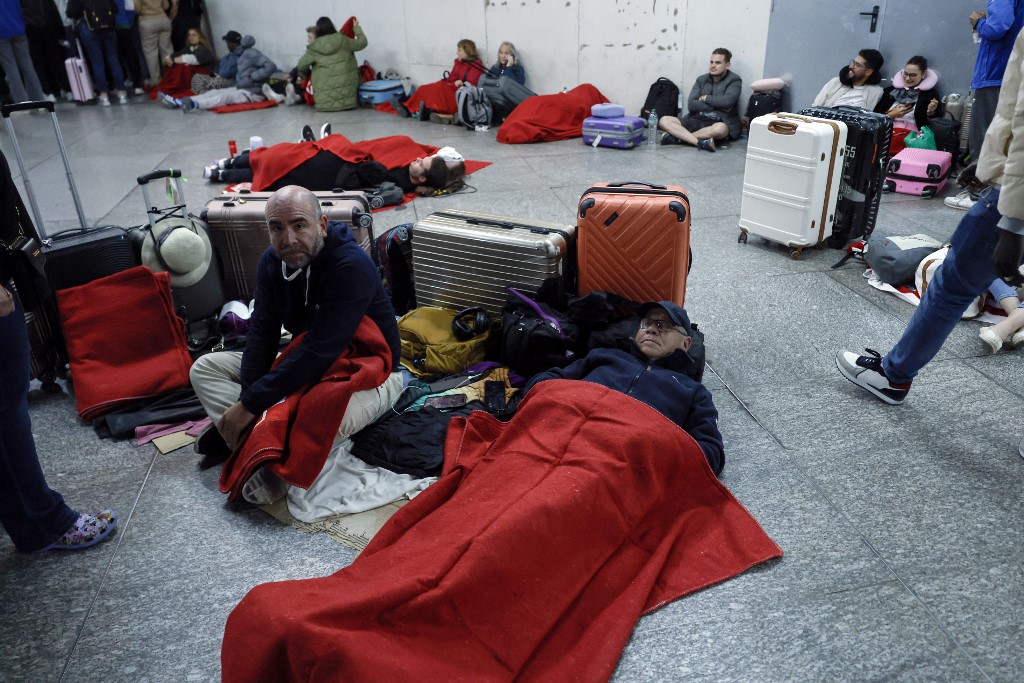 Des personnes allongées près de leurs valises dans une gare de Madrid. Ils ont des couvertures rouges pour se réchauffer.