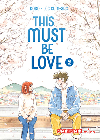 Couverture du livre 'This Must Be Love 2' montrant deux jeunes gens assis sur un banc sous des cerisiers en fleurs, avec une ville et des collines en arrière-plan.