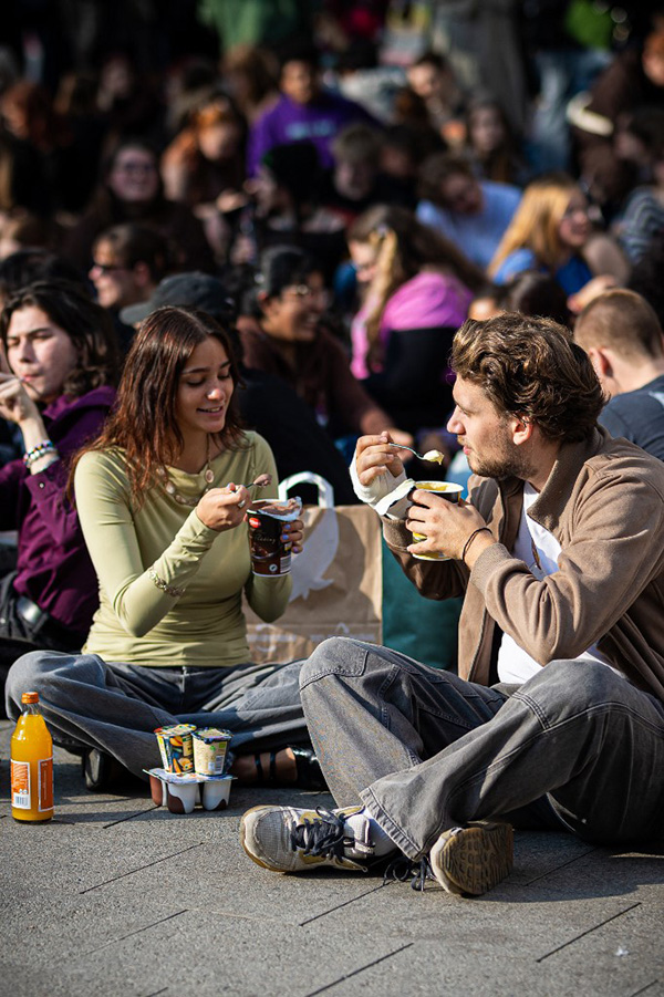 photo de jeunes adultes assis à même le sol sur une place et mangeant leurs desserts à la fourchettes. On voit qu'ils sont nombreux