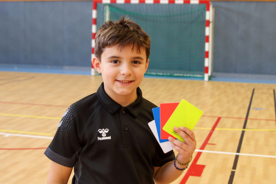 Gauthier, sur un terrain de handball, montre les 4 cartons de couleurs (blanc, bleu, rouge et jaune) qu'il utilise en tant qu'arbitre.