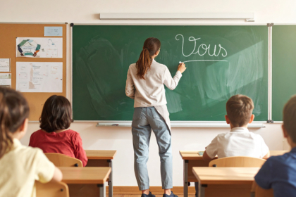 une enseignante écrit le mot "Vous" au tableau devant des élèves assis à leurs places.
